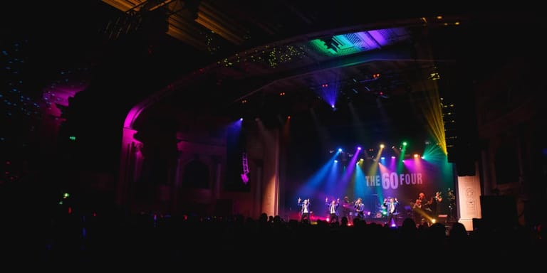 Multiple performers on stage bathed in vibrant blue, pink and purple stage lighting during a live concert performance.