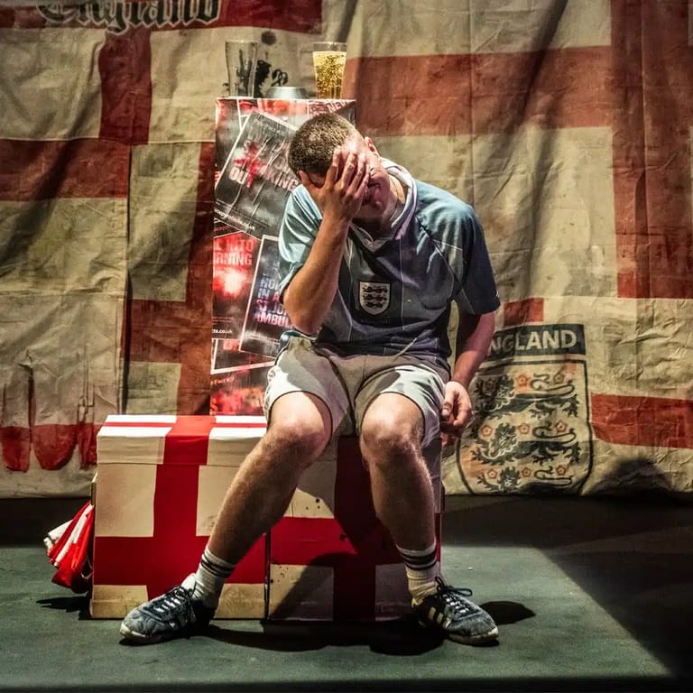 Performer in England football kit holding head against large Union Jack flag backdrop during Fringe show.