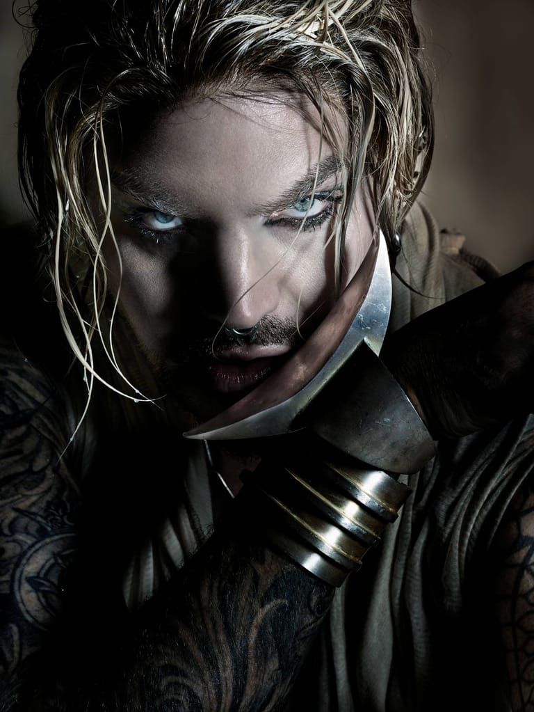 Person with platinum-blonde streaked hair and bold eye makeup, wearing black leather and metallic neck rings, gazes downward in dramatic stage lighting.