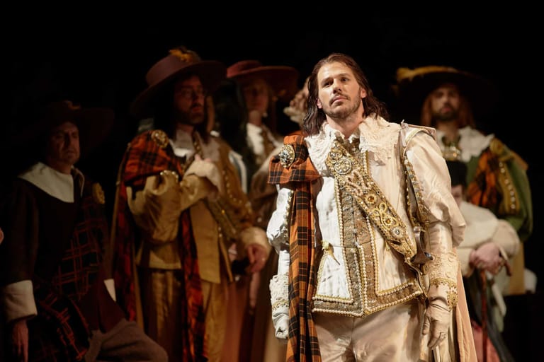 Actor in ornate period costume and cloak stands centre-stage amid costumed ensemble in theatrical production.
