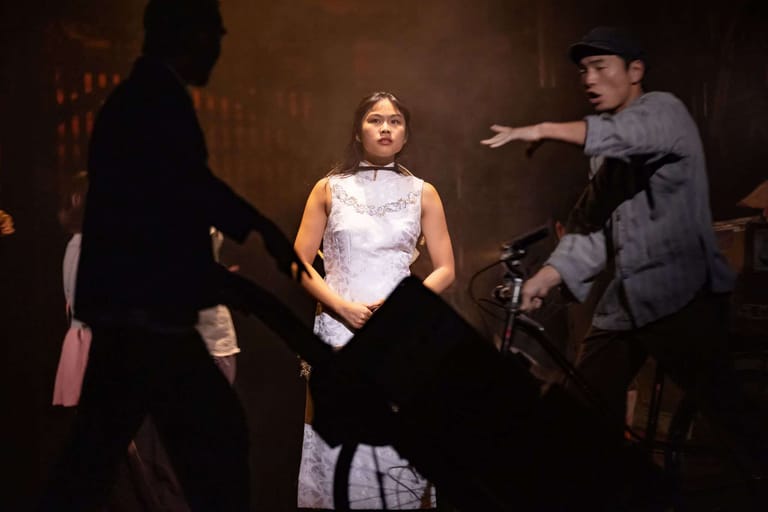 Actress in a white sleeveless top surrounded by male performers in formal attire on stage.