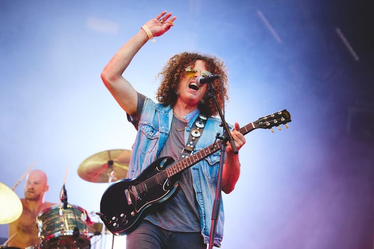 Curly-haired guitarist in denim shirt and sunglasses raises fist while performing with electric guitar on stage.