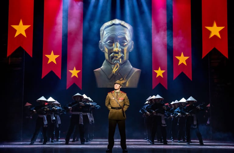 Performer in gold suit stands centre stage beneath projected portrait, flanked by musicians in red-lit setting with Vietnamese flags.