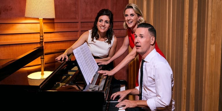 Three musicians collaborating at a grand piano in a warmly lit studio setting.