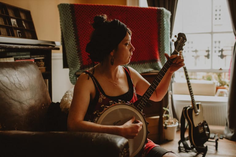 Musician in a studio holding a stringed instrument, with a red backdrop and guitars visible nearby.