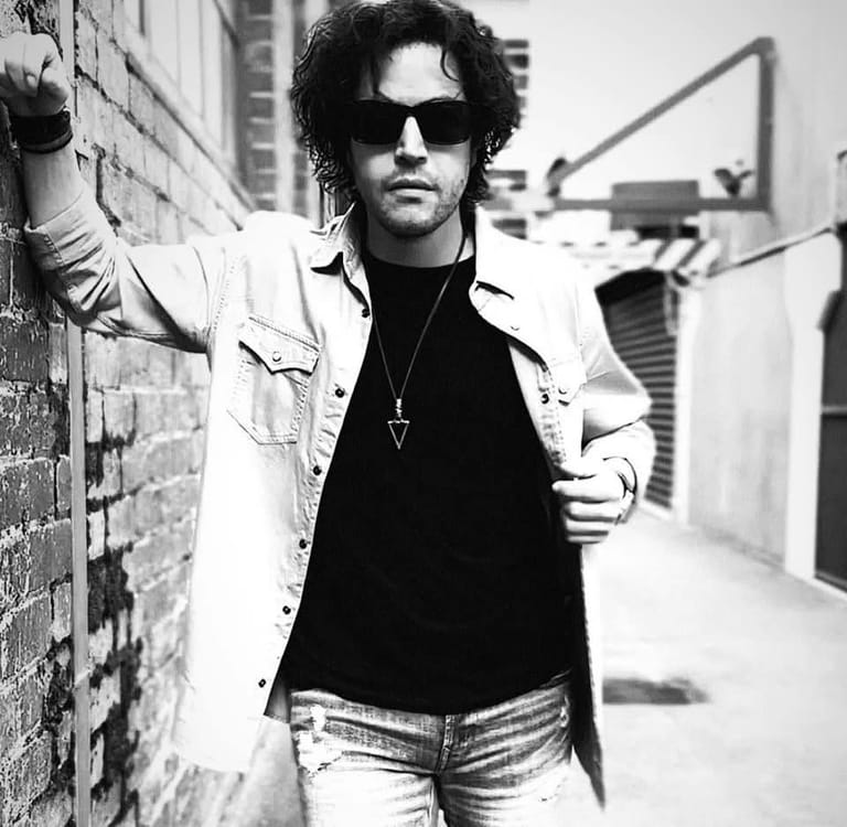 Man in sunglasses and white jacket leans against brick wall in monochrome urban setting.