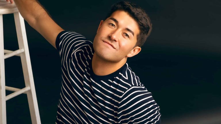 Performer in a striped shirt poses against a dark teal background beside a white ladder.