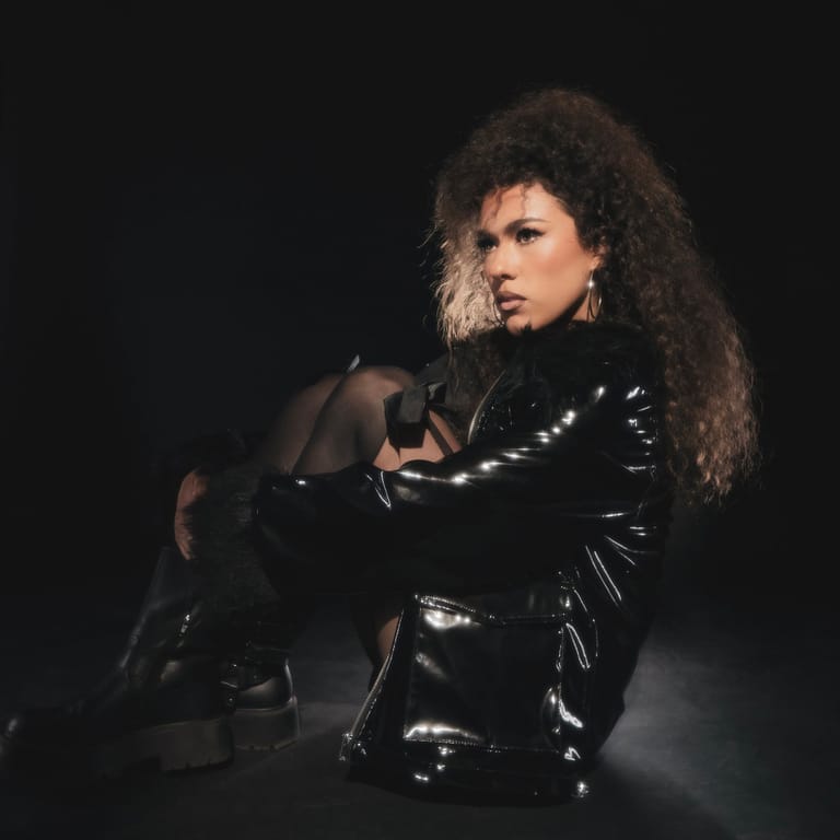 Performer with voluminous curled hair wearing a glossy black jacket against a dark background.
