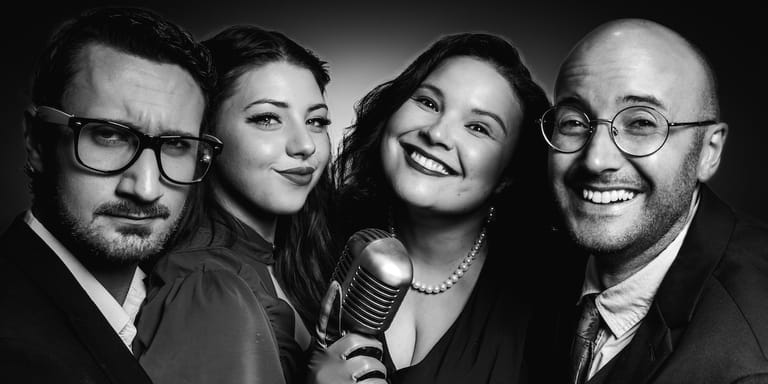 Four performers in formal attire pose together against a dark background, with one holding a vintage microphone.