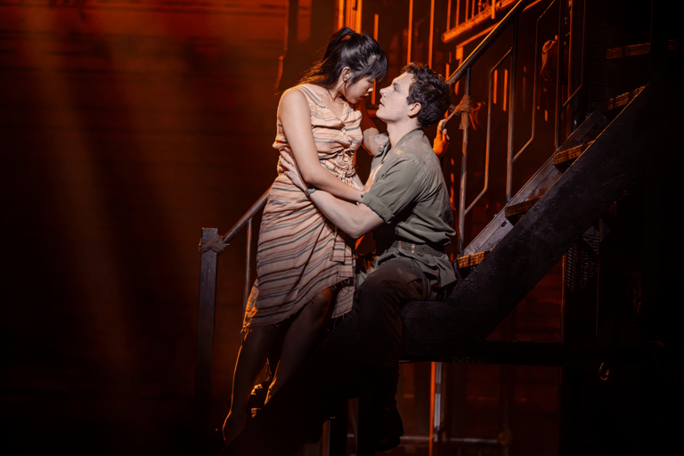 Two performers embrace on a dimly lit wooden staircase during an intimate theatre scene.