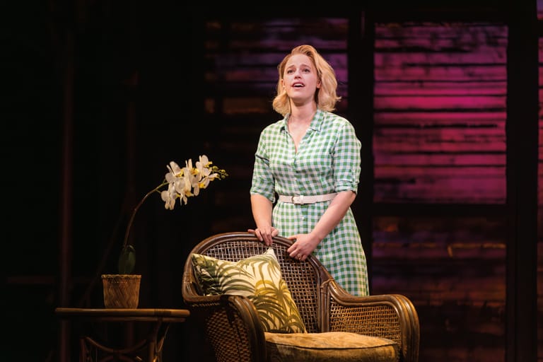 Woman in gingham dress stands beside wicker furniture and dried flowers on dimly lit stage.
