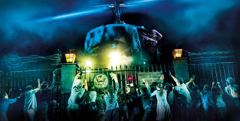 Helicopter descends towards a crowd of people reaching upward in a dramatic blue-lit scene.