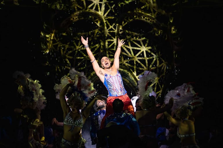 Performer elevated above ensemble cast with arms raised in triumph against illuminated golden branch set design.
