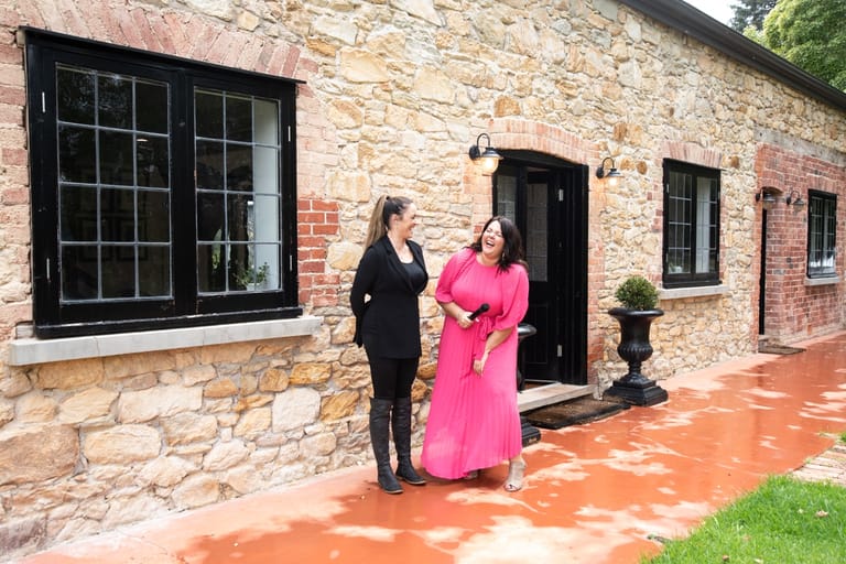 Two women stand outside a heritage brick building, one wearing black and one in a bright pink dress.