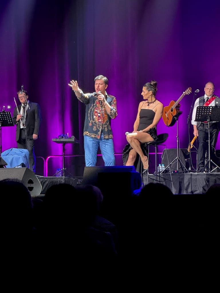 Singer in patterned shirt gestures to audience whilst performing with band members under purple stage lighting.
