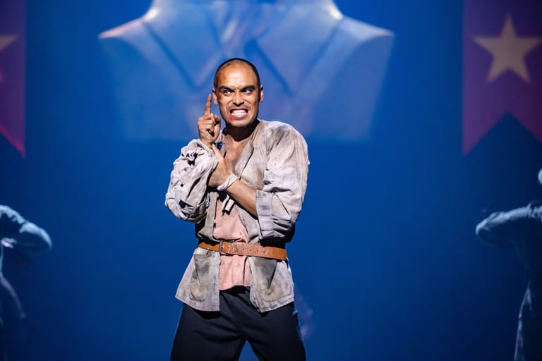 Man in white shirt and pink belt performs on stage with microphone against blue starlit backdrop.