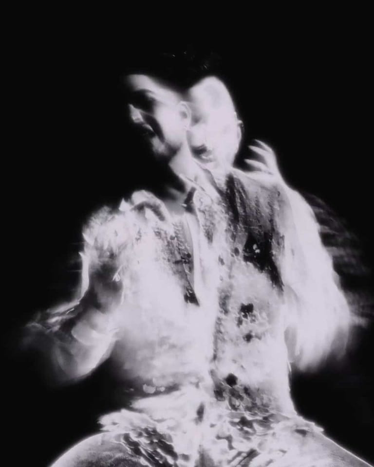 Performer in textured garment with arms raised against dark background, captured in high-contrast black and white.