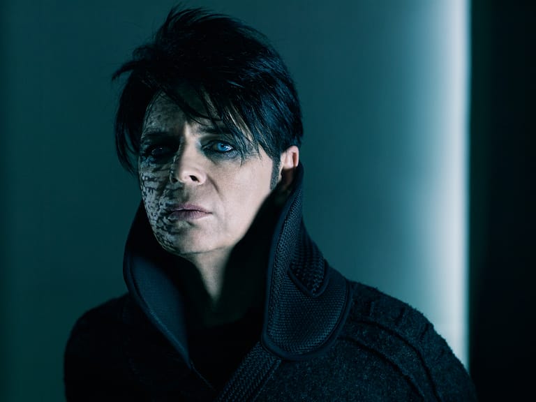 Person with dark hair and facial glitter against a teal-lit backdrop, wearing a black textured jacket.