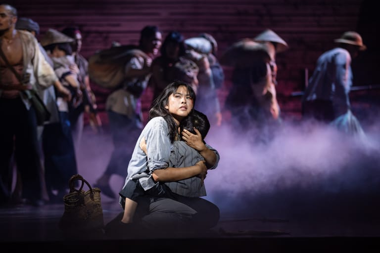 Performer kneeling centre stage amid purple-lit fog, surrounded by costumed ensemble cast members.