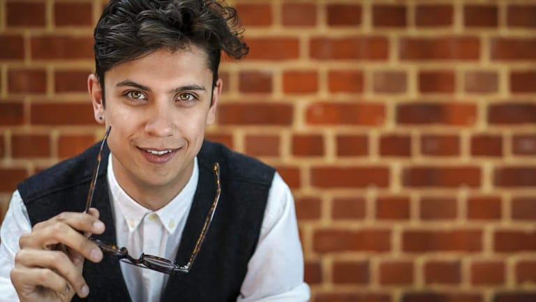 Young man in black vest and white shirt holding sunglasses, smiling against a brick wall.