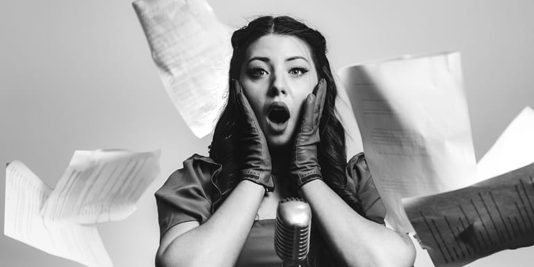 Singer with hands to face expressing surprise, surrounded by white papers in black and white photograph.