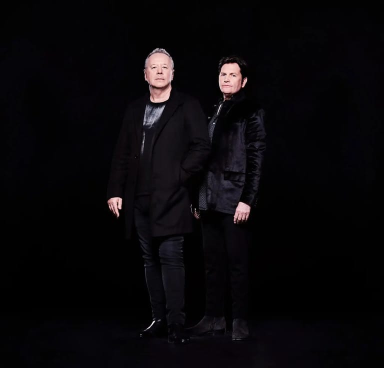 Two performers in dark clothing stand together against a black background during a stage performance.