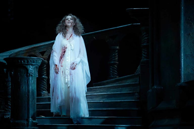 Performer in flowing white gown stands on dimly lit stage stairs under blue theatrical lighting.