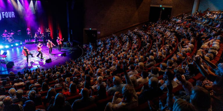 Packed crowd of concert-goers watching performers on a brightly lit stage in a large venue.