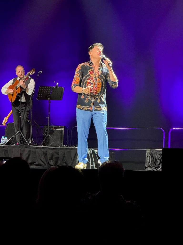 Singer in patterned shirt performs at microphone with guitarist on stage under purple lighting.