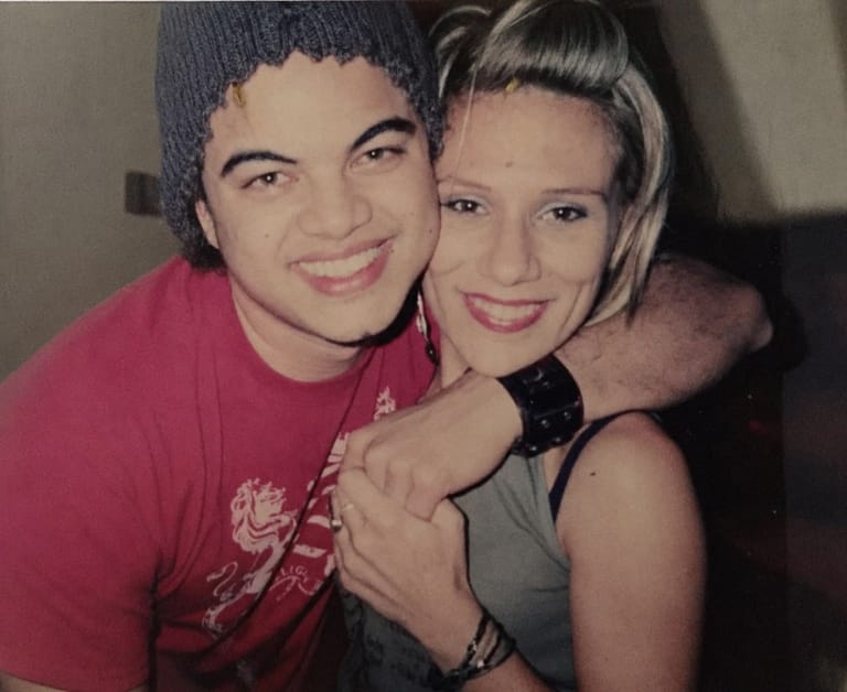 Two young people smiling at the camera, one wearing a pink shirt and grey beanie, the other in a grey tank top.