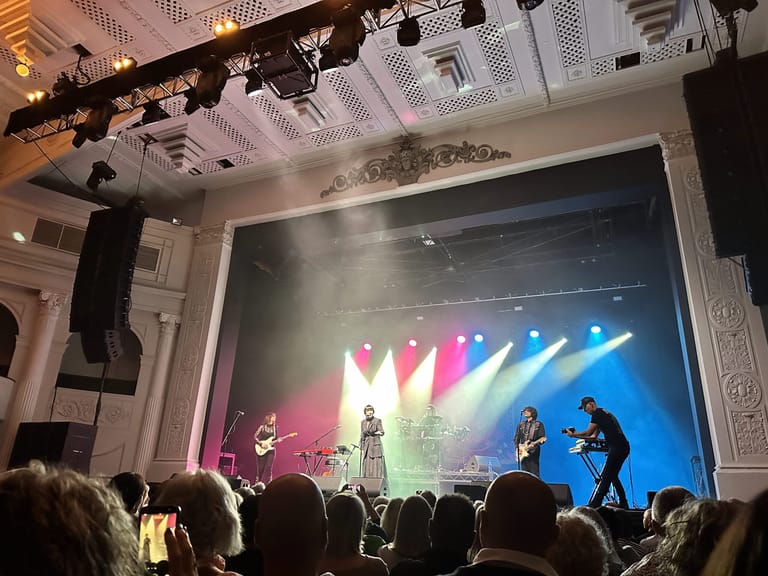 Singer in centre stage spotlights during theatre performance, silhouetted audience in foreground.
