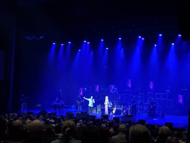 Singer in cream suit performs at centre stage under intense blue spotlights with full band and audience in foreground.