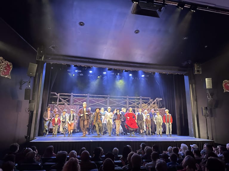 Large cast ensemble in period costumes takes a final bow on stage under blue theatrical lighting.