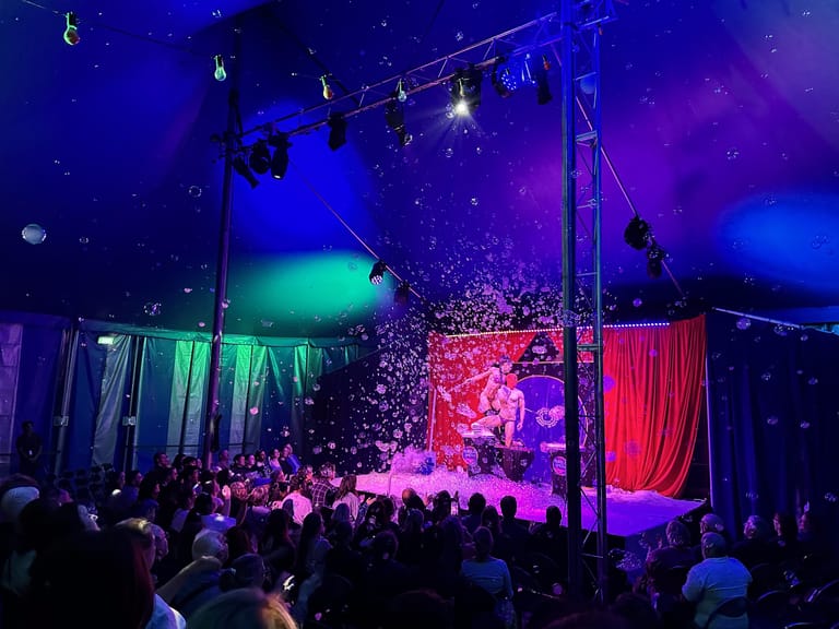 Colourful bubbles cascade across a dimly lit theatre stage surrounded by an audience, illuminated by vibrant blue, pink and green stage lighting.