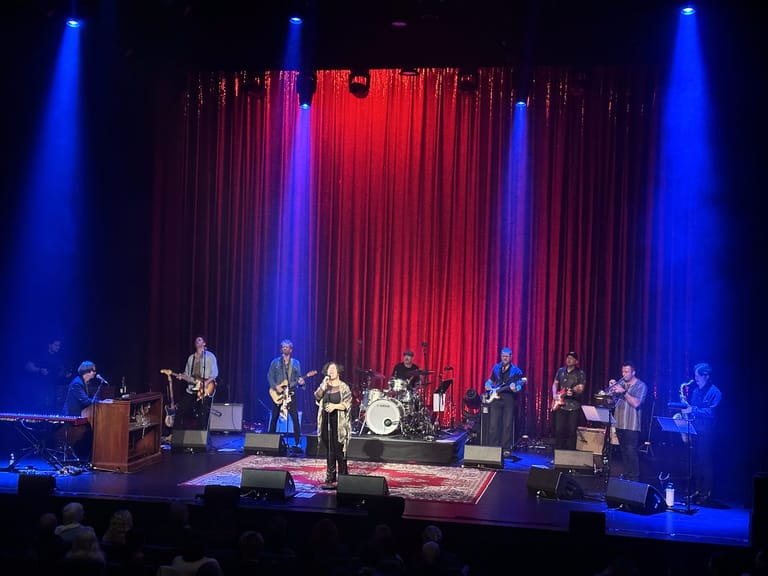Full band and singers performing on stage with red velvet curtains lit by blue and red spotlights.