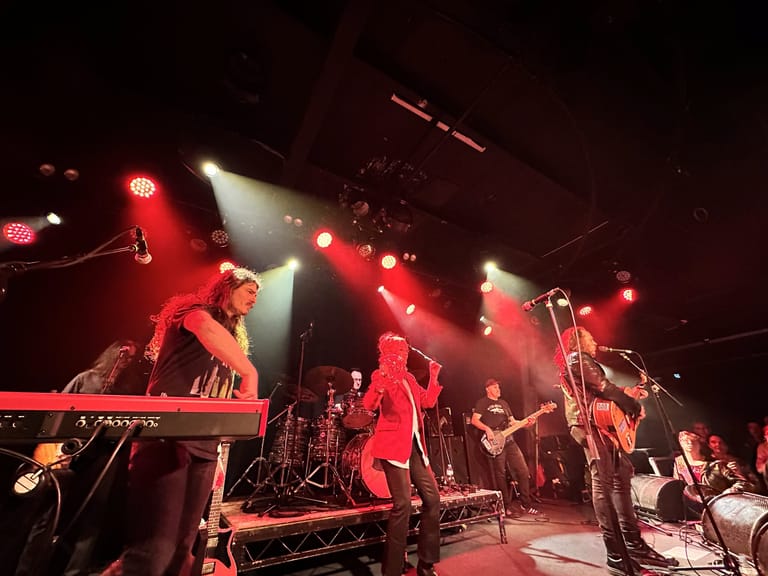 Five performers on stage bathed in red and white spotlights during a live band performance.