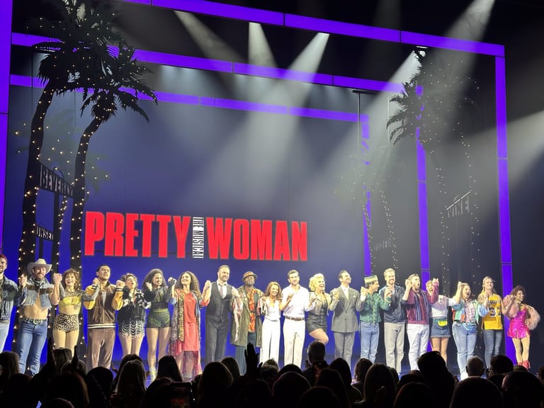 Cast members stand on stage beneath "Pretty Woman" title, lit by purple spotlights and a palm tree set piece.