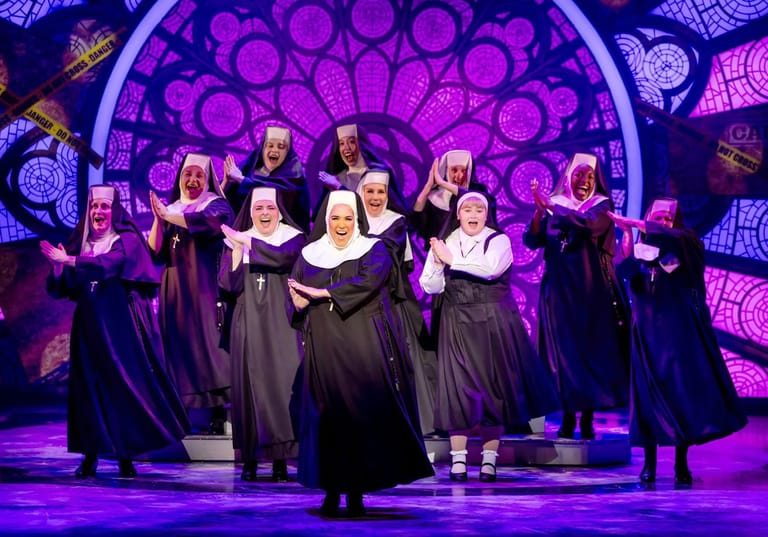 Choir in dark robes performing on stage beneath purple circular light installation.