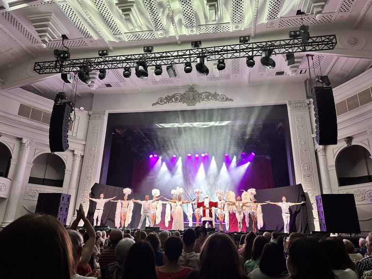 Ensemble of performers in festive costumes on stage beneath purple spotlights during a Christmas Spectacular production.