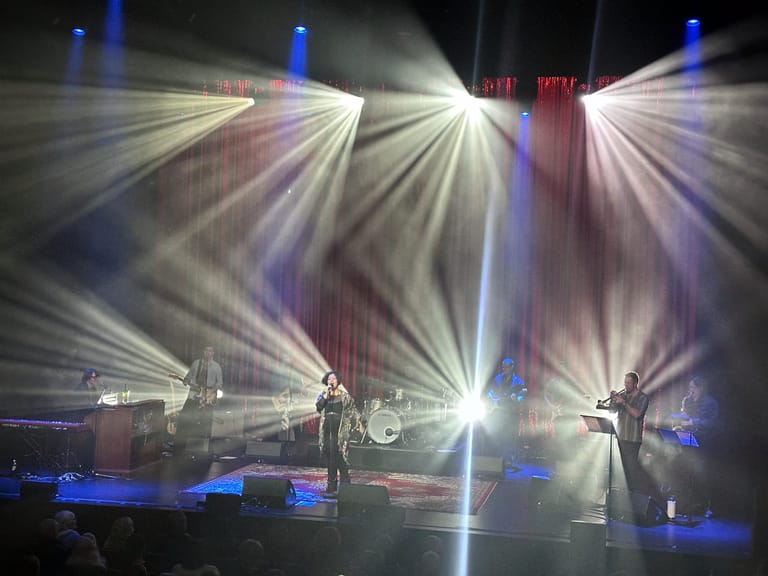 Two performers on stage bathed in dramatic blue and white spotlight beams radiating outward.