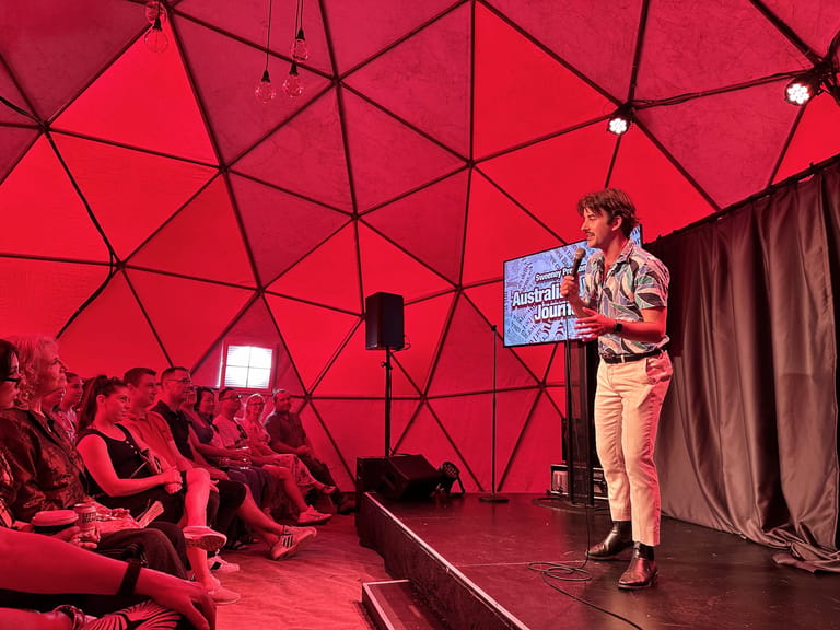 Performer stands on stage holding a newspaper prop under red geometric dome lighting, addressing seated audience members.