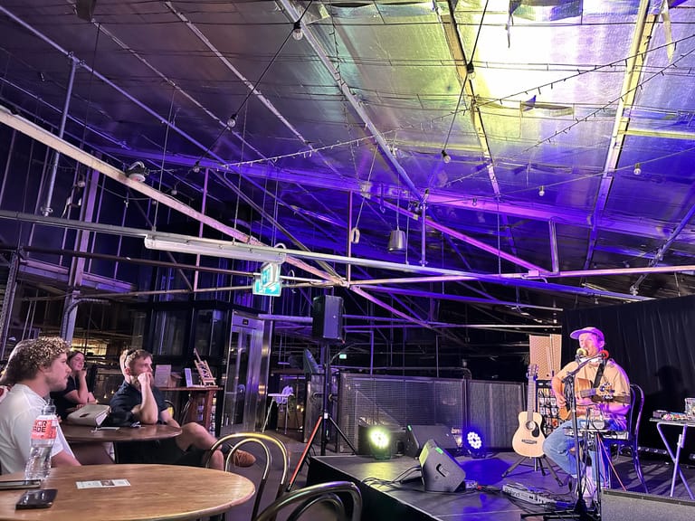 Musician performing with acoustic guitar on stage under purple lighting while audience members watch from seated tables.