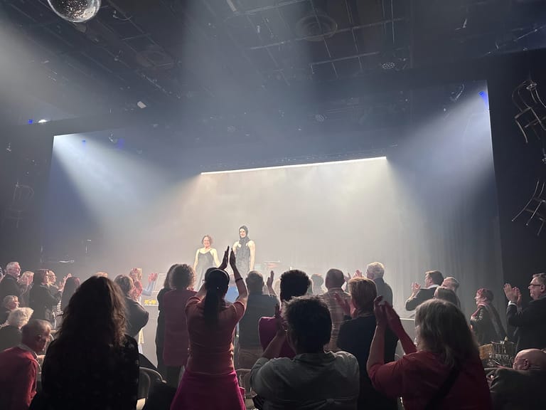 Two performers stand centre stage beneath dramatic spotlights whilst an audience surrounds them on a thrust stage.