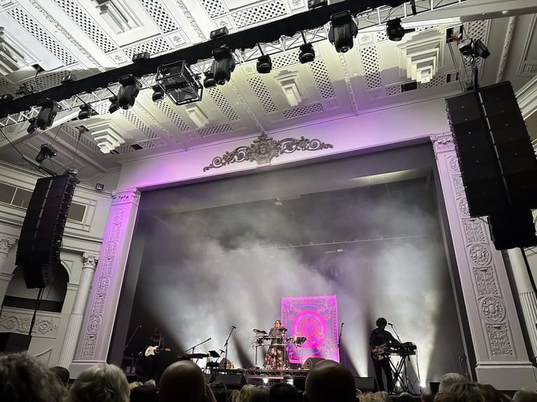 Stage performance with purple lighting, large projection screen, and silhouetted audience members in foreground.
