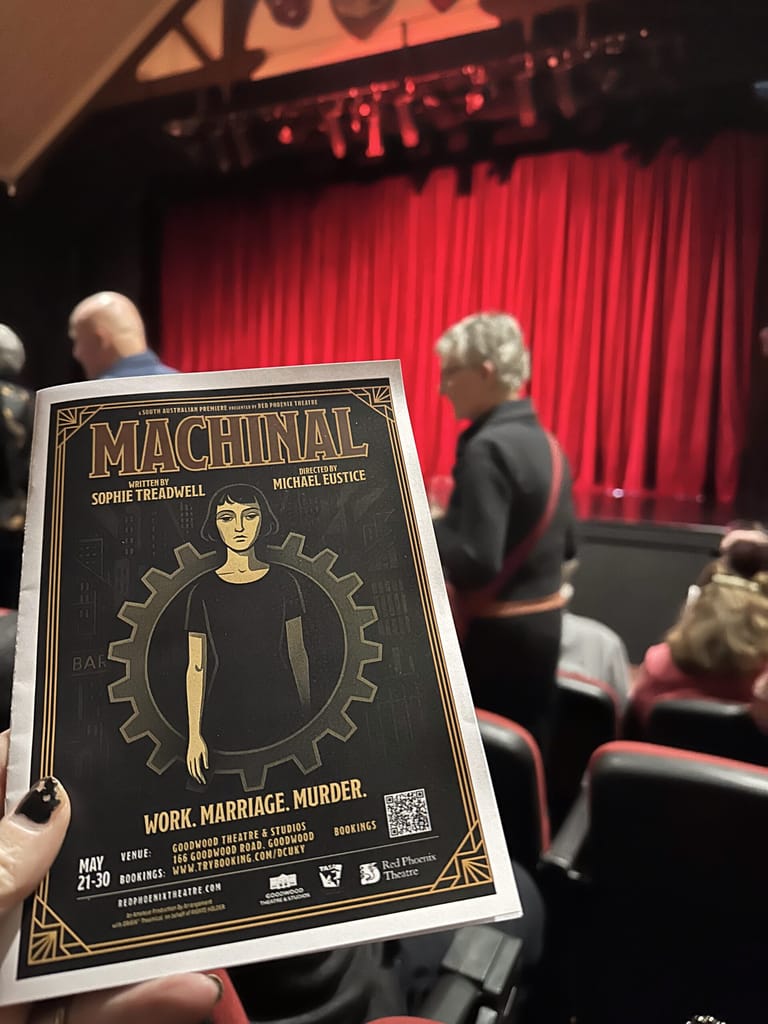Hand holding a Machinal theatre programme featuring Art Deco-style design with gears and a figure, red stage curtains visible behind.