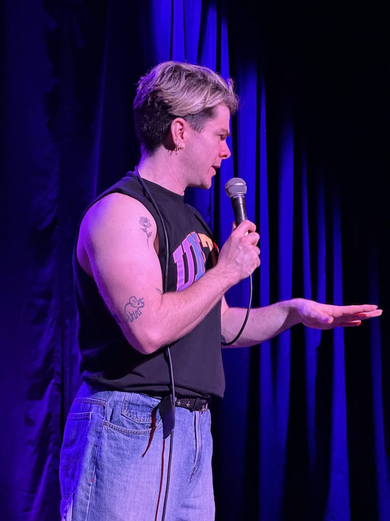 Performer on stage holding a microphone, lit by blue spotlights, wearing a black sleeveless shirt and jeans.