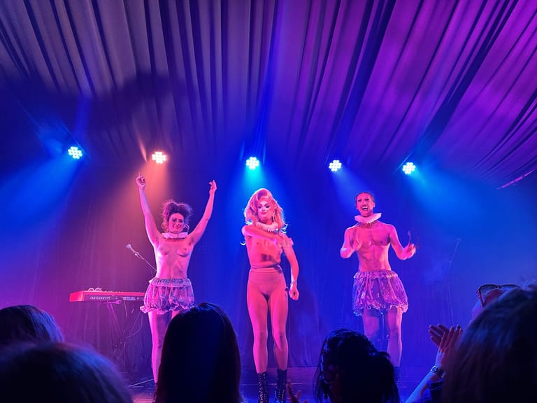 Three performers in sequined outfits dance under blue and pink stage lights during a cabaret show.