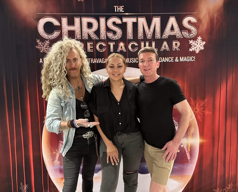 Three performers pose together in front of The Christmas Spectacular promotional banner with red and gold stage lighting.