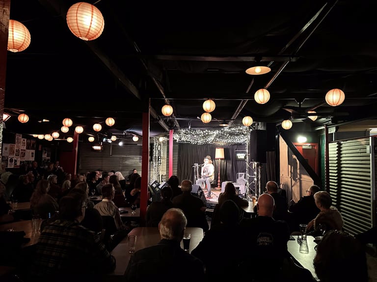 Audience members gathered in an intimate venue lit by paper lanterns, watching a performance on stage in the distance.