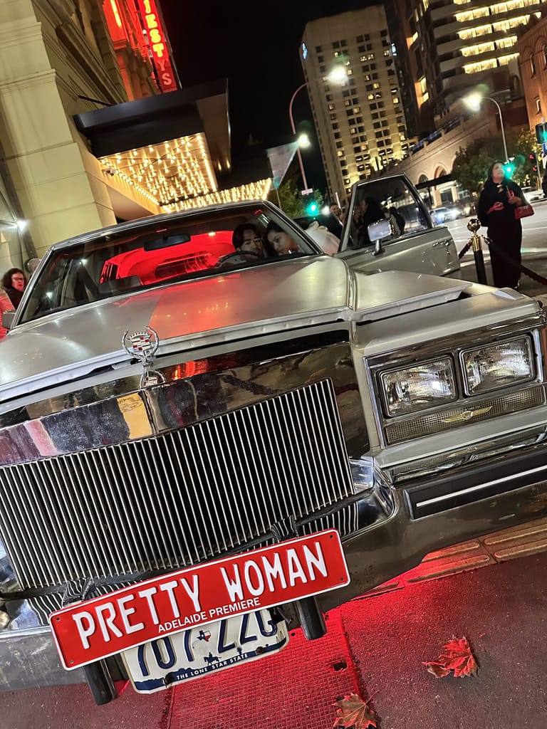 Classic silver convertible with "Pretty Woman" number plate parked on an illuminated city street at night.